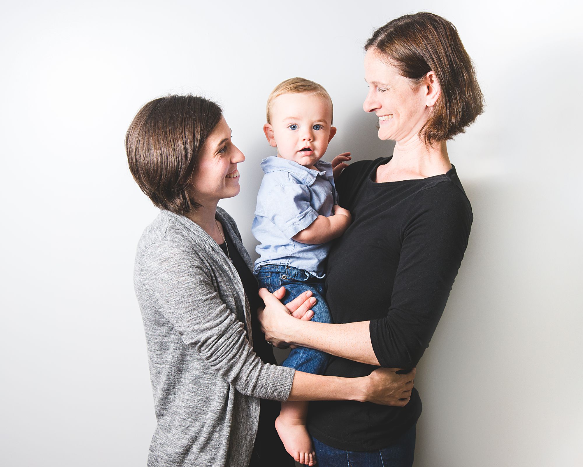 Two moms in grey neutral clothes with their baby boy in blue jeans in casual studio portrait