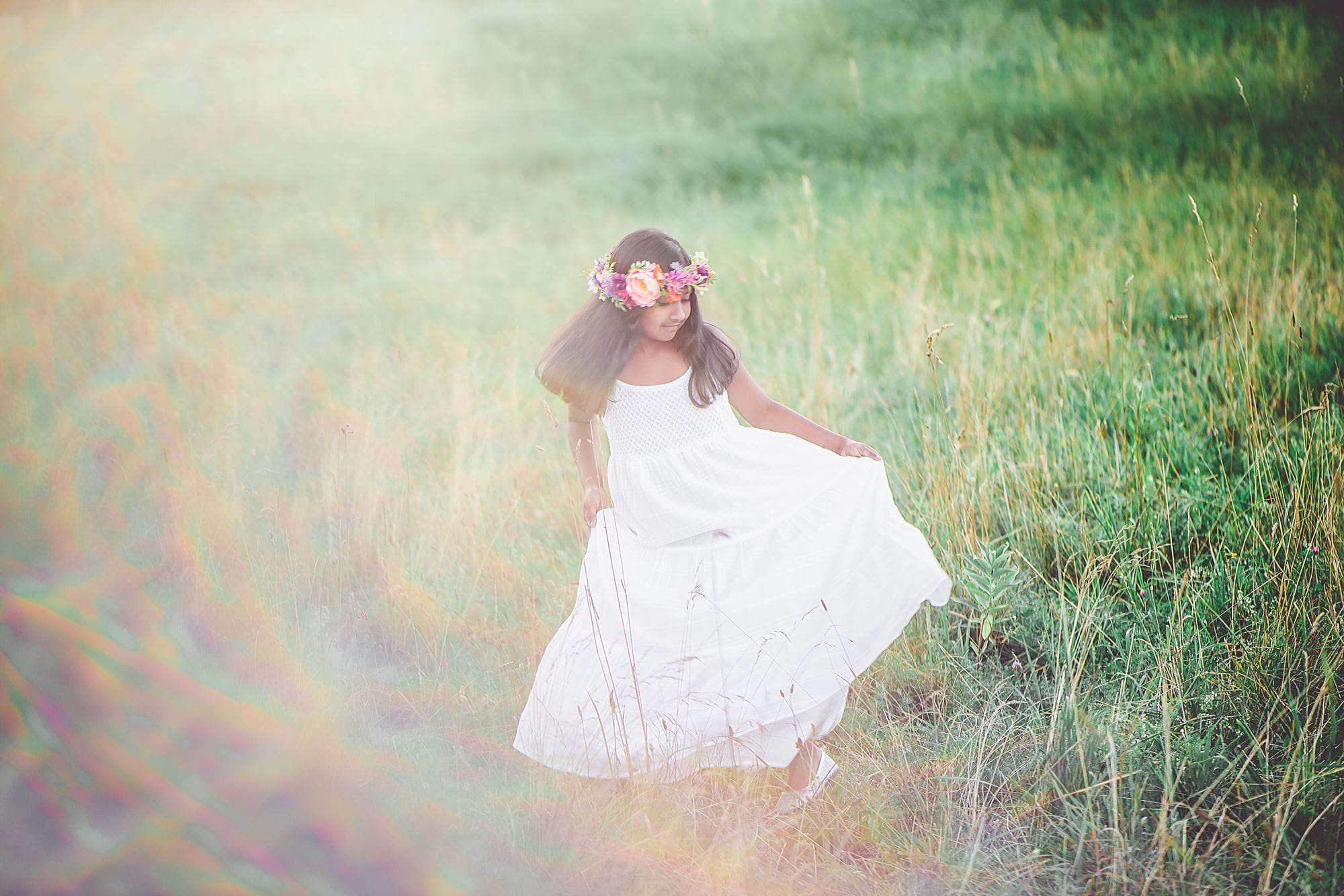 Girl in long white dress and flower crown outside in a grassy meadow with sunflare