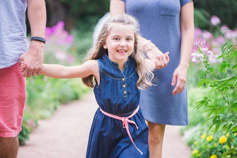 Close up of little girl holding hands with her parents walking on a path, wearing a blue dress with pink belt